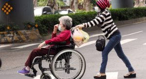 Masked caregiver pushes masked lady in wheelchair